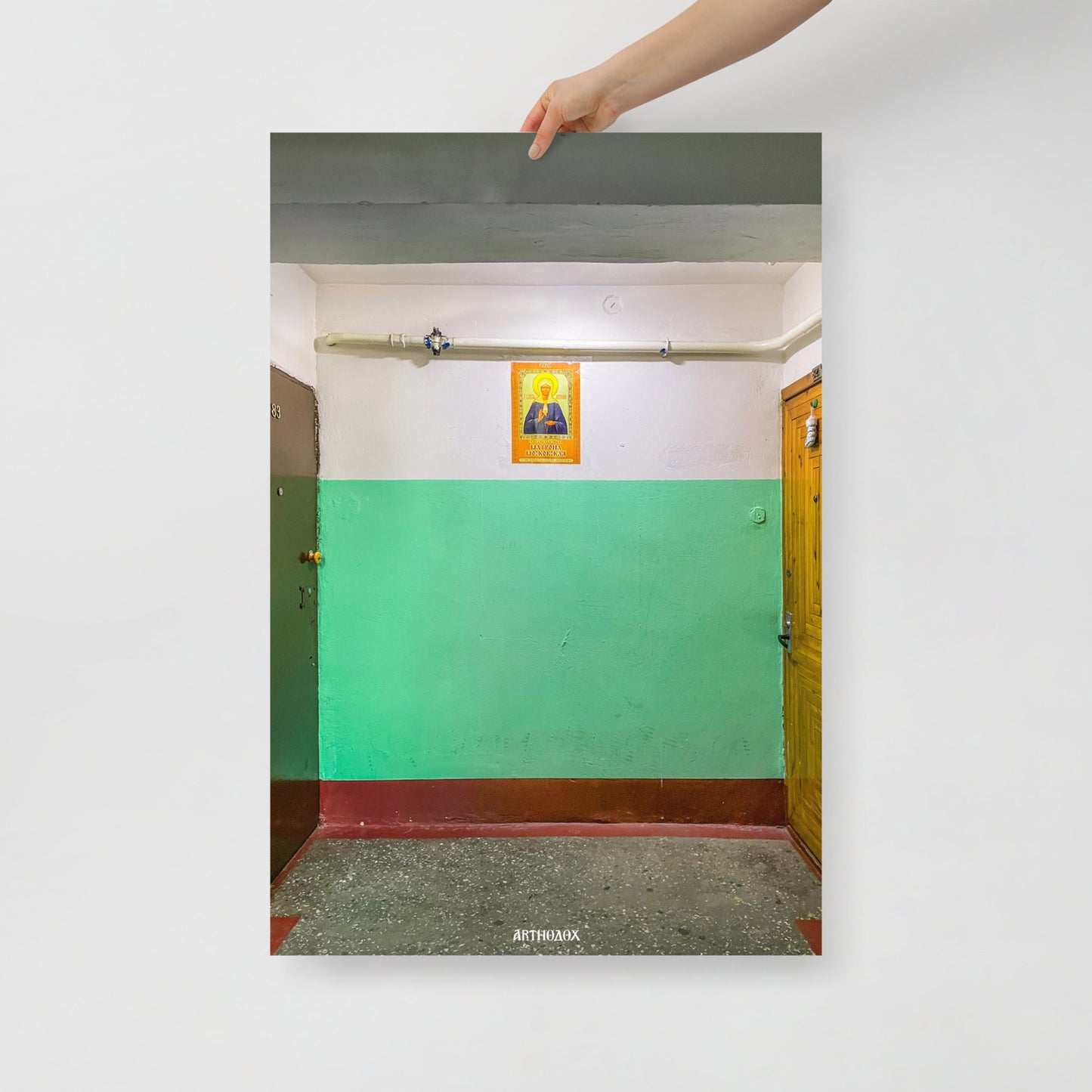 Hand holding large 91x61cm print depicting a typical Russian apartment stairwell featuring an icon of Holy Blessed Matrona of Moscow on the wall. From arthodox.com