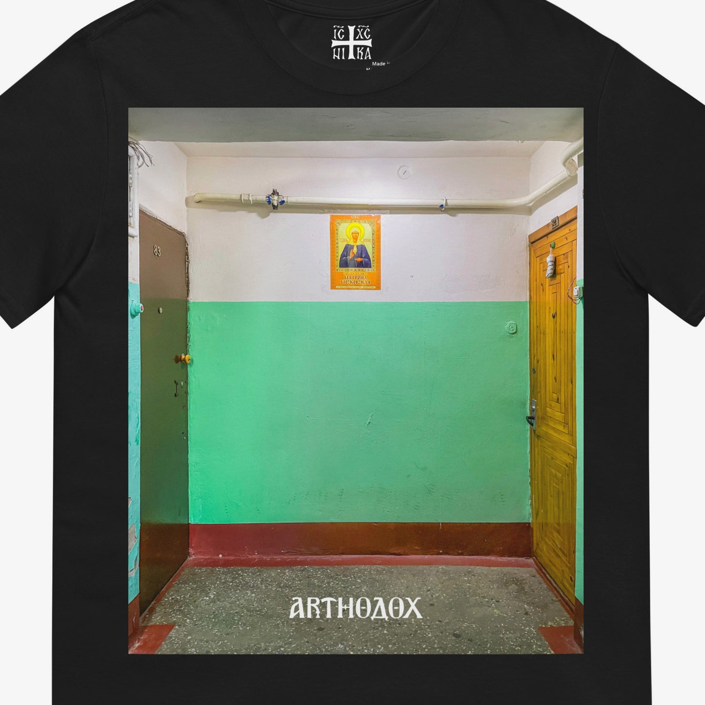 Black unisex T-shirt with a large print depicting a typical Russian apartment stairwell featuring an icon of Holy Blessed Matrona of Moscow on the wall. Closeup photo. From arthodox.com